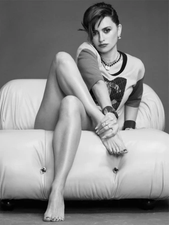 Monochrome portrait of Penelope Cruz seated on a tufted leather sofa wearing a t-shirt and layered necklaces styled by Helga Burrill