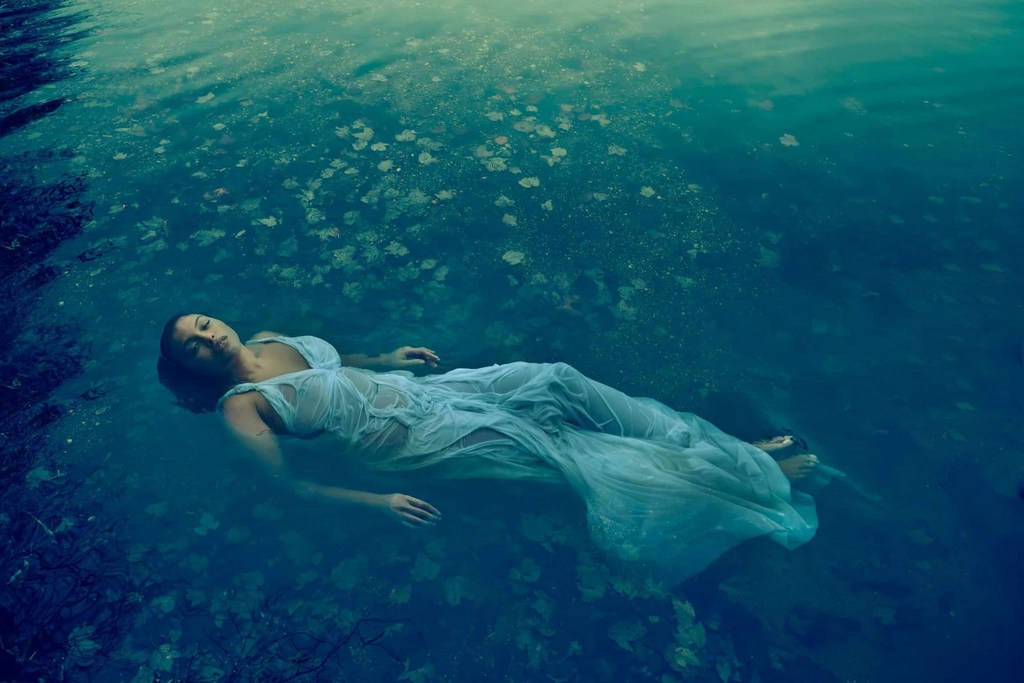 Woman in a sheer white dress floats in dark blue water with dappled sunlight and scattered leaves, creating a tranquil and ethereal scene.
