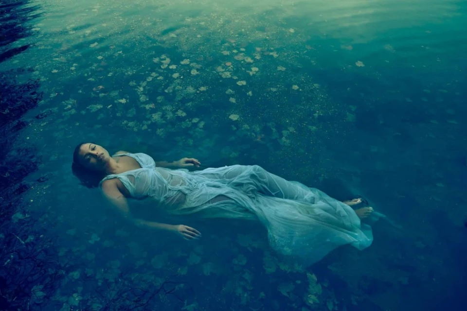 Woman in a sheer white dress floats in dark blue water with dappled sunlight and scattered leaves, creating a tranquil and ethereal scene.
