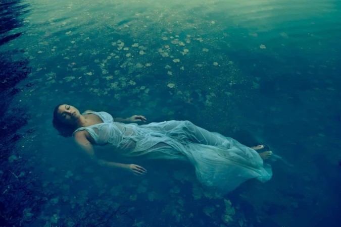 Woman floating on her back in a dark turquoise water body wearing a wet white semi-sheer dress.