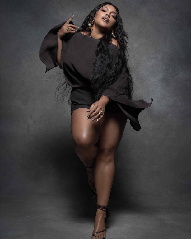 Portrait of a plus-size woman with long, dark hair, wearing a dark, off-the-shoulder dress and high-heeled sandals in a studio shot.