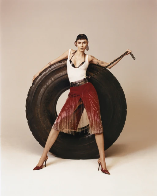 A female fashion model poses in front of a large black tire wearing a white tank top, a red beaded skirt, and matching red heels.