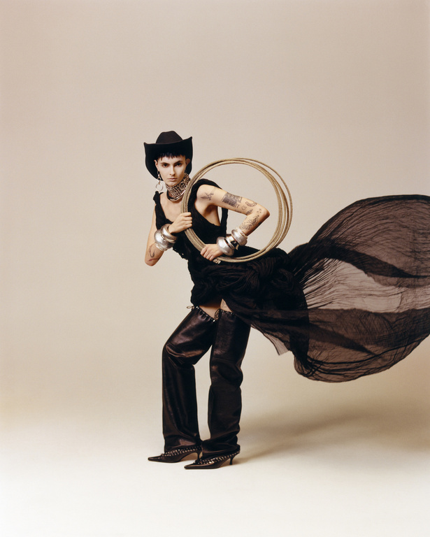 Woman in black cowboy hat and dress holds circular hula hoops, posing in a studio setting. Full-length shot with neutral background.