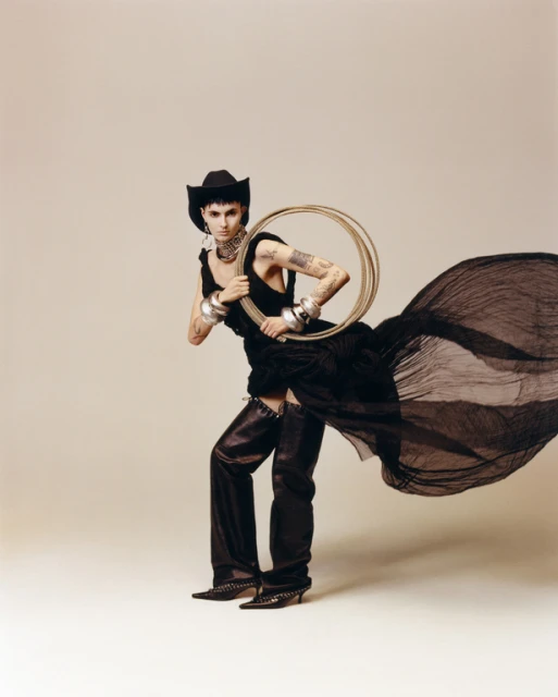 Woman in black cowboy hat and dress holds circular hula hoops, posing in a studio setting. Full-length shot with neutral background.