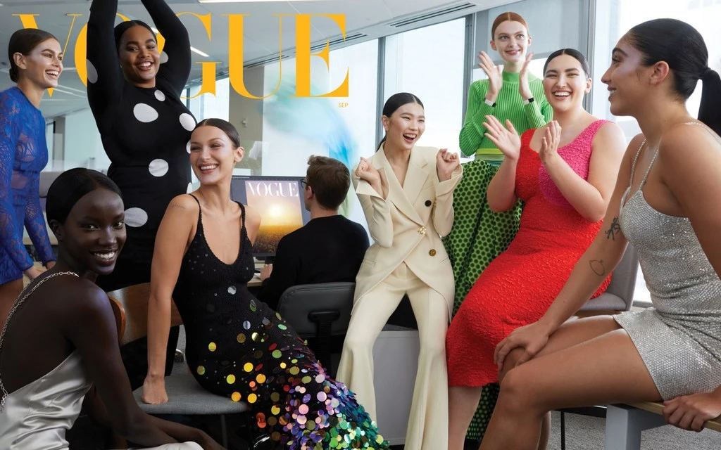 Fashion editorial shot featuring a diverse group of nine smiling fashion models in various dresses and a pantsuit, posed in an office setting.