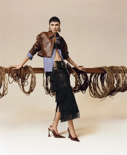 A stylish woman in a brown jacket and black fringe skirt poses indoors, complemented by neutral tones and wrapped rope accents.