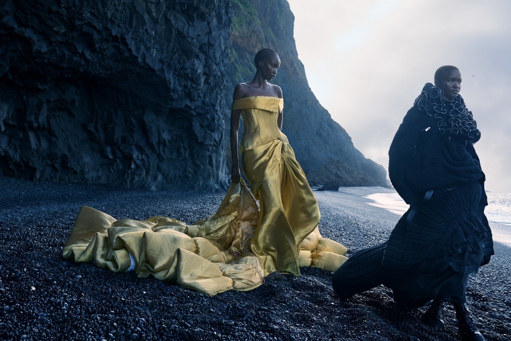 Two women in elegant gowns pose on a black sand beach with a dramatic cliff backdrop, creating a striking fashion editorial image.