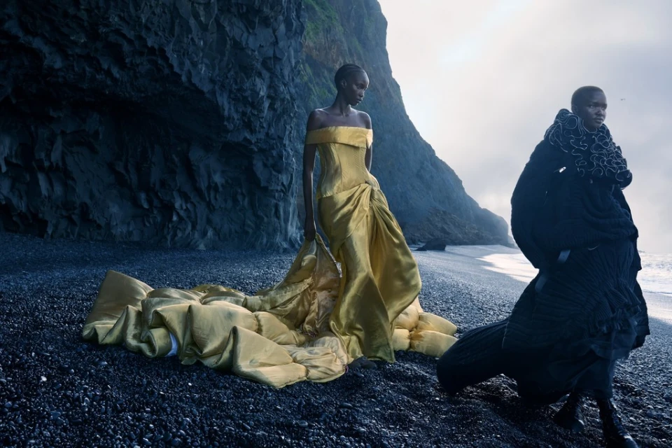 Two women in elegant gowns pose on a black sand beach with a dramatic cliff backdrop, creating a striking fashion editorial image.