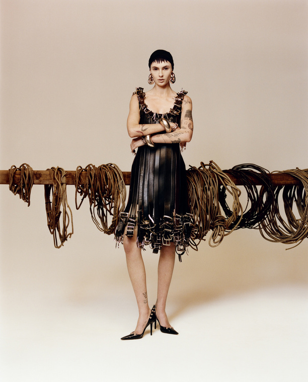 Fashion model poses in a black leather dress, jewelry, and black heels in front of a wood beam with hanging belts and ropes.