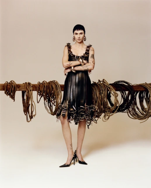 Fashion model poses in a black leather dress, jewelry, and black heels in front of a wood beam with hanging belts and ropes.