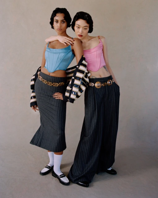 Two women in 90s fashion pose in studio. One woman in a blue corset and skirt places her arm on another in a pink corset and trousers.