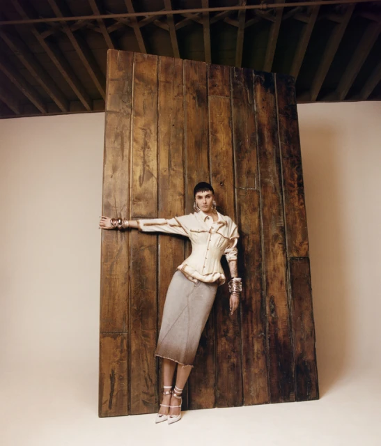Adult female model posing while leaning against a wooden plank in a photo studio, wearing a beige top, grey skirt and heels.