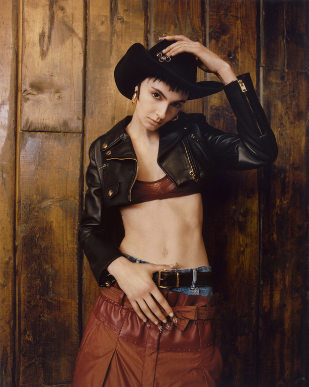 Woman in cowboy hat, cropped black leather jacket, and red leather skirt posing against a textured wooden wall in an indoor setting.