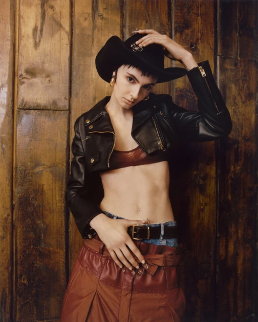 Woman in cowboy hat, cropped black leather jacket, and red leather skirt posing against a textured wooden wall in an indoor setting.