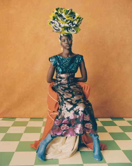 A fashionable Black woman seated in an orange-toned studio wears a floral dress, blue boots, and a large, colorful floral headpiece.