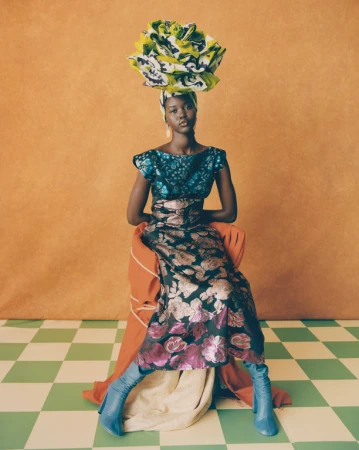 A fashionable Black woman seated in an orange-toned studio wears a floral dress, blue boots, and a large, colorful floral headpiece.