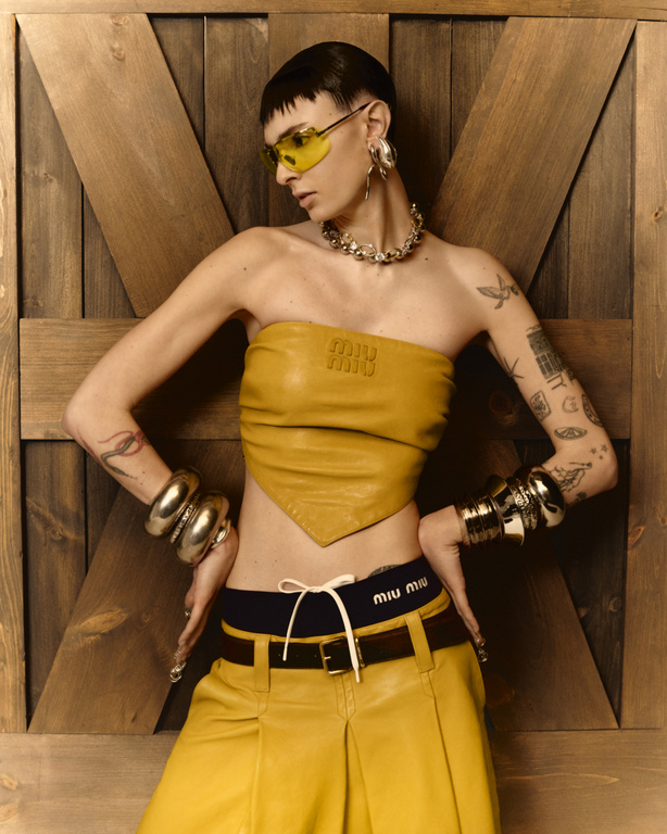 A slender person with short hair, sunglasses, and jewelry poses in a yellow top and pants against a wooden backdrop.