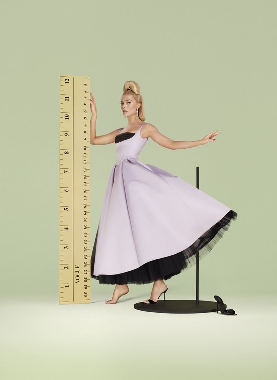 Woman in a pastel dress poses next to an oversized ruler in a studio. The shot is brightly lit with a pale green background.