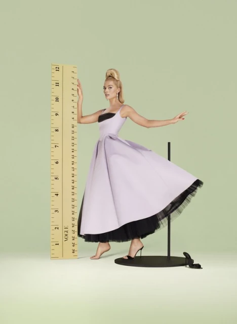 Woman in a pastel dress poses next to an oversized ruler in a studio. The shot is brightly lit with a pale green background.