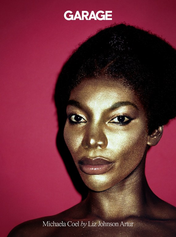 Close-up portrait of Michaela Coel with golden skin and dark hair against a vibrant pink background; bold beauty statement.
