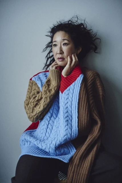 Portrait of Sandra Oh wearing a multi-colored knit sweater against a neutral wall background in a softly lit, indoor environment.