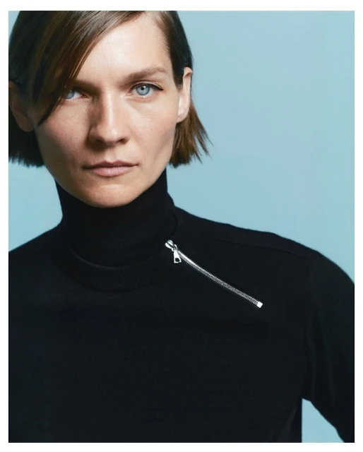 Close-up studio shot of an adult woman with short brown hair wearing a black turtleneck sweater with a silver zipper on a light blue background.