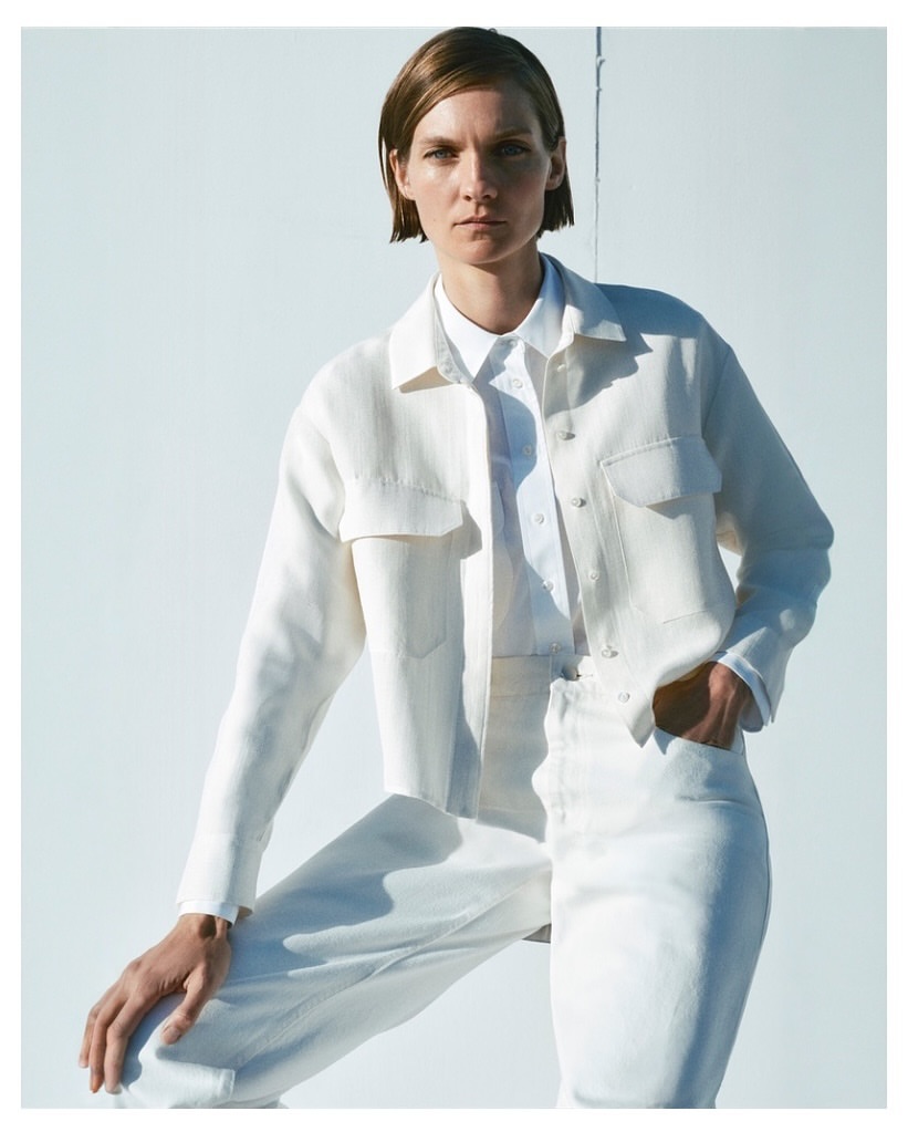 Model with short brown hair wearing white shirt and white jacket against a bright white background
