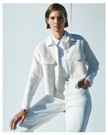 Model with short brown hair wearing white shirt and white jacket against a bright white background