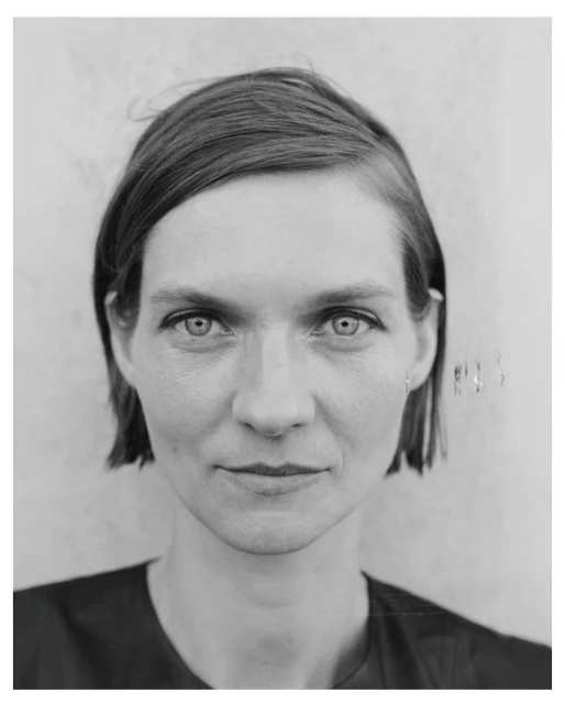 Monochrome portrait of a fair-skinned adult woman with short brown hair, facing forward in a dark top against a mottled grey background.
