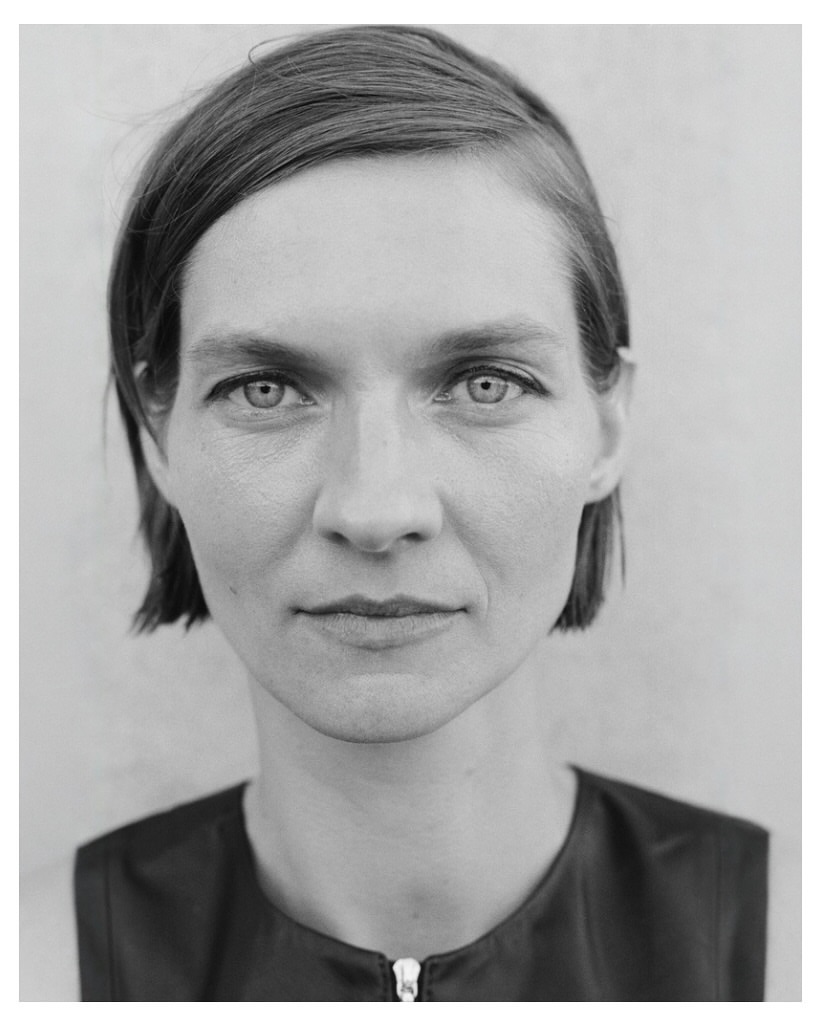 Close-up black and white portrait of a woman with short hair and a zippered dark top against a neutral wall.