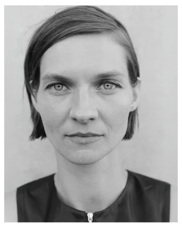Close-up black and white portrait of a woman with short hair and a zippered dark top against a neutral wall.