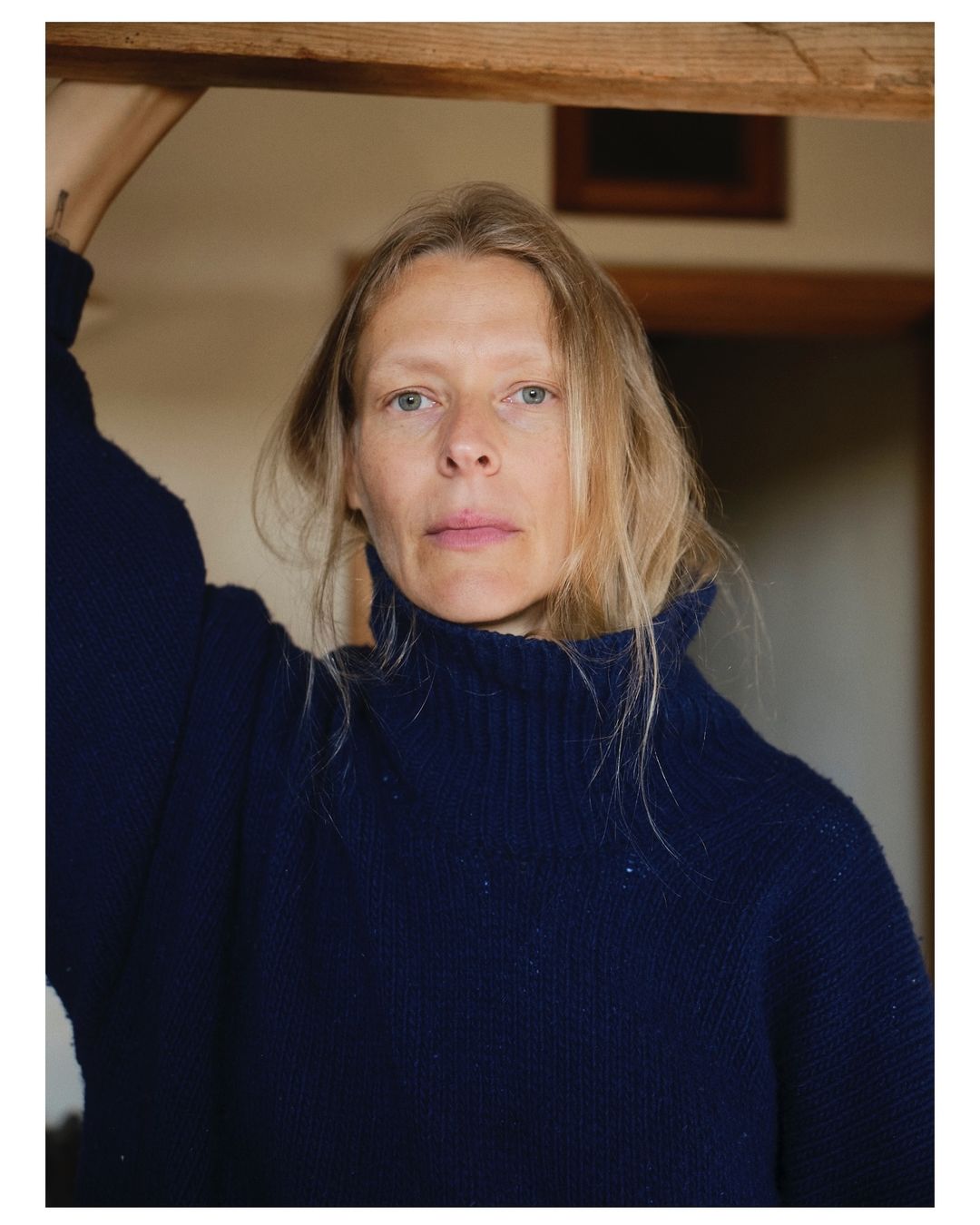 Blonde woman in a blue turtleneck sweater stands indoors with neutral expression and fair skin in soft lighting. Wooden frame visible.