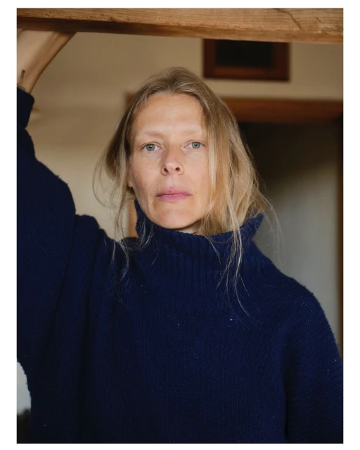 Blonde woman in a blue turtleneck sweater stands indoors with neutral expression and fair skin in soft lighting. Wooden frame visible.