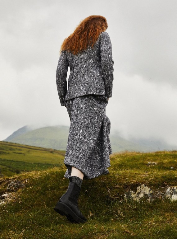 Woman in a gray tweed suit walks on a grassy hill with mountains in the background under a cloudy sky, fashion editorial shot.