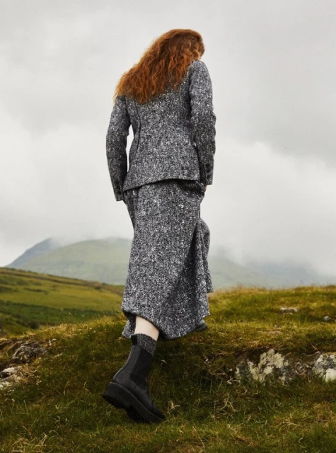 Woman in a gray tweed suit walks on a grassy hill with mountains in the background under a cloudy sky, fashion editorial shot.