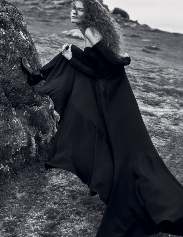 Monochrome portrait of a woman in a long black gown posing on a rocky hillside, her curly hair blowing in the wind.