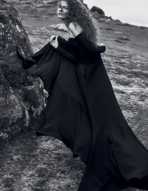 Monochrome portrait of a woman in a long black gown posing on a rocky hillside, her curly hair blowing in the wind.