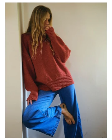 Woman leaning against a plain wall wearing an oversized rust red sweater and blue denim trousers