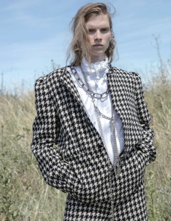 A woman stands in a field wearing a houndstooth suit, white blouse, and chain necklaces against a blue sky and grassy backdrop.