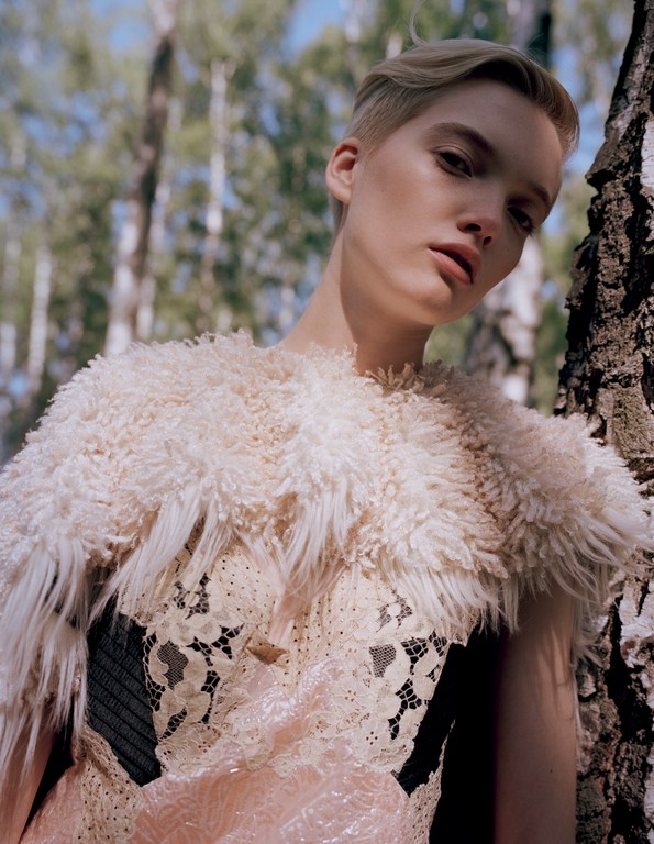 Portrait of a blonde woman with short hair in a lace top with a fuzzy overlay, posing outdoors in front of birch trees in a forest.