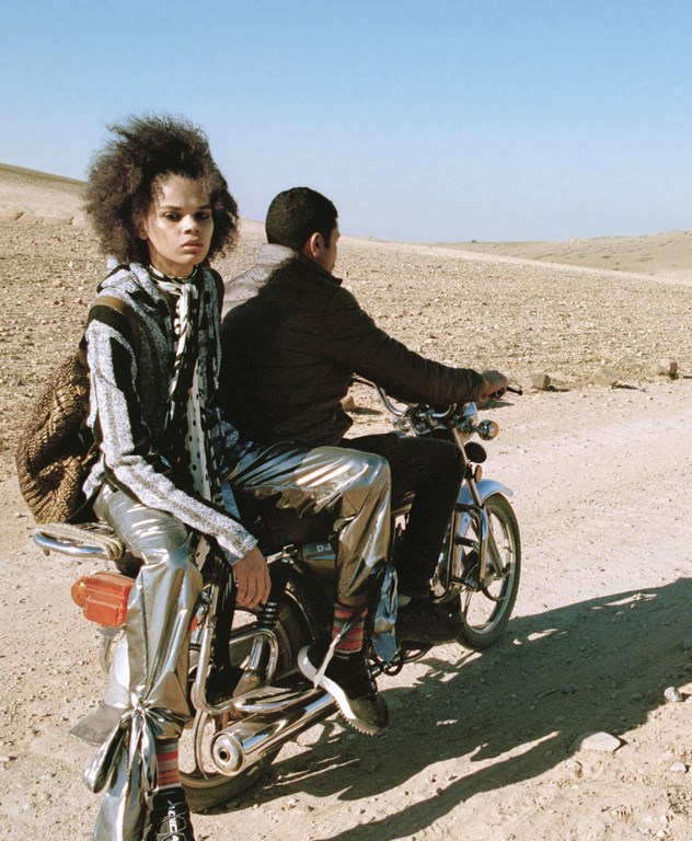 Two people on a motorcycle in a desert landscape, one with an afro and metallic pants, under a clear sky. Fashion editorial.