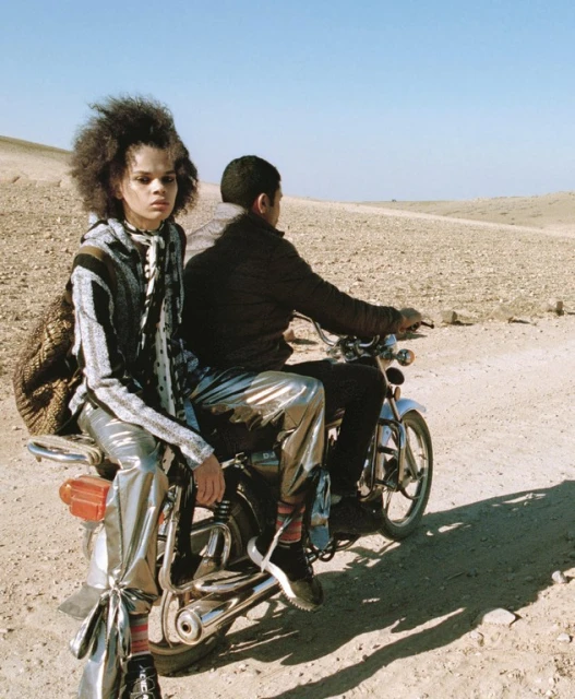 Two people on a motorcycle in a desert landscape, one with an afro and metallic pants, under a clear sky. Fashion editorial.
