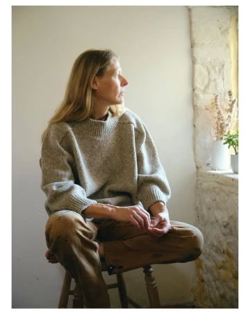 Woman with long blonde hair wearing a chunky knit sweater and brown trousers, seated on a wooden stool indoors.