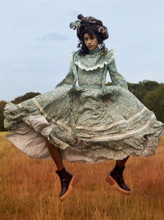 Woman in a full floral dress and platform boots appears to float above a golden grassy field, with a cloudy sky and trees.