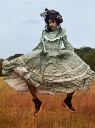 Woman in a full floral dress and platform boots appears to float above a golden grassy field, with a cloudy sky and trees.