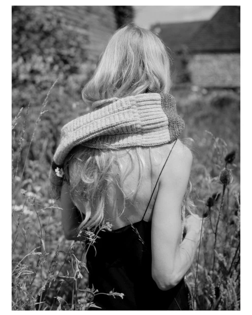 Back view of a woman with long blonde hair wearing a chunky knit scarf, standing in a field of tall grass with a building in the distance.