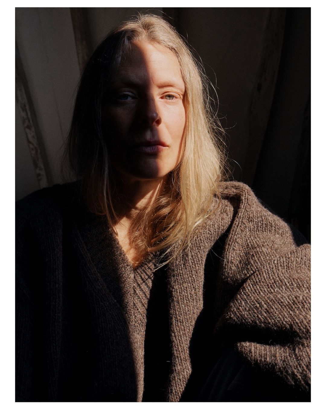 Portrait of a fair-skinned woman with shoulder-length blonde hair wearing a brown knitted sweater, dramatically lit by sunlight.
