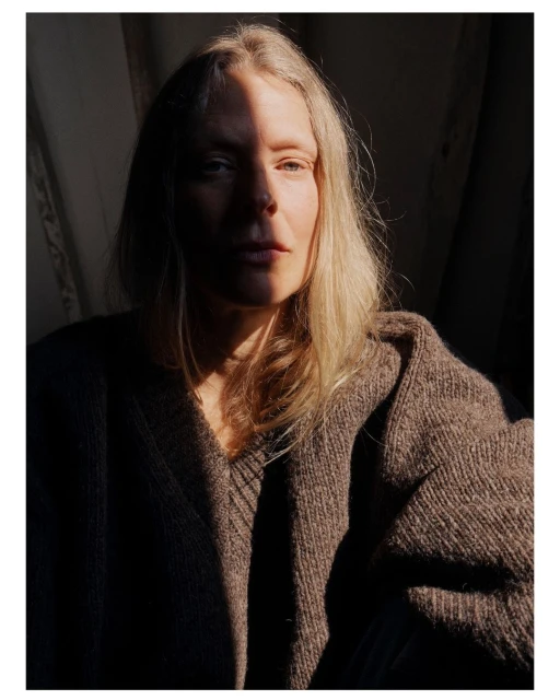 Portrait of a fair-skinned woman with shoulder-length blonde hair wearing a brown knitted sweater, dramatically lit by sunlight.