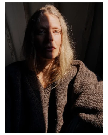 Portrait of a fair-skinned woman with shoulder-length blonde hair wearing a brown knitted sweater, dramatically lit by sunlight.
