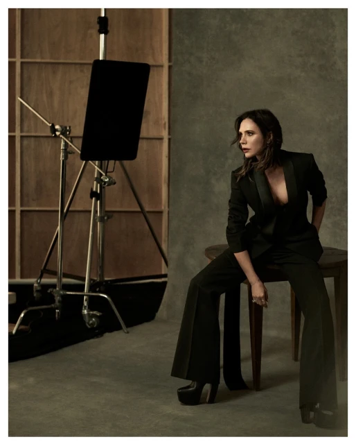 Candid studio portrait of a stylish adult woman in a black suit, seated on a stool, with studio lighting and neutral backdrop.